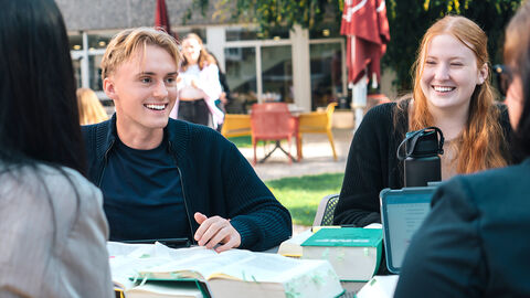 Studienzentrum Ein Anwärter und eine Anwärterin lächeln während des Lernens draußen im Innenhof der Hochschule.